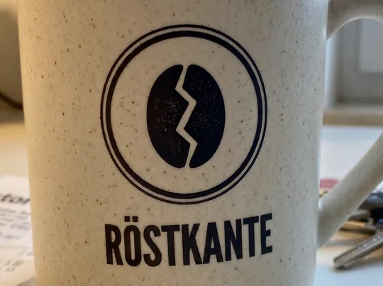 Handyfoto in Nahaufnahme: das Logo mit Doppelring, Kaffeebohne mit Zickzack-Linie und dem Schriftzug Röstkante