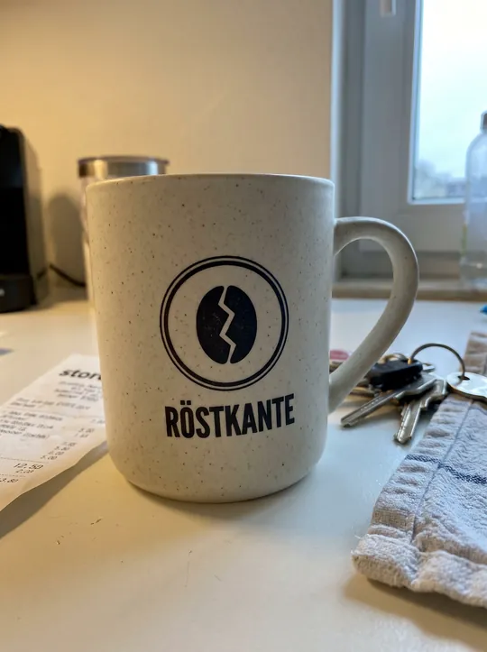Handyfoto: weißer gesprenkelter Becher mit Röstkante-Logo auf einer Küchentheke neben Schlüsseln, Kassenbon und Geschirrtuch
